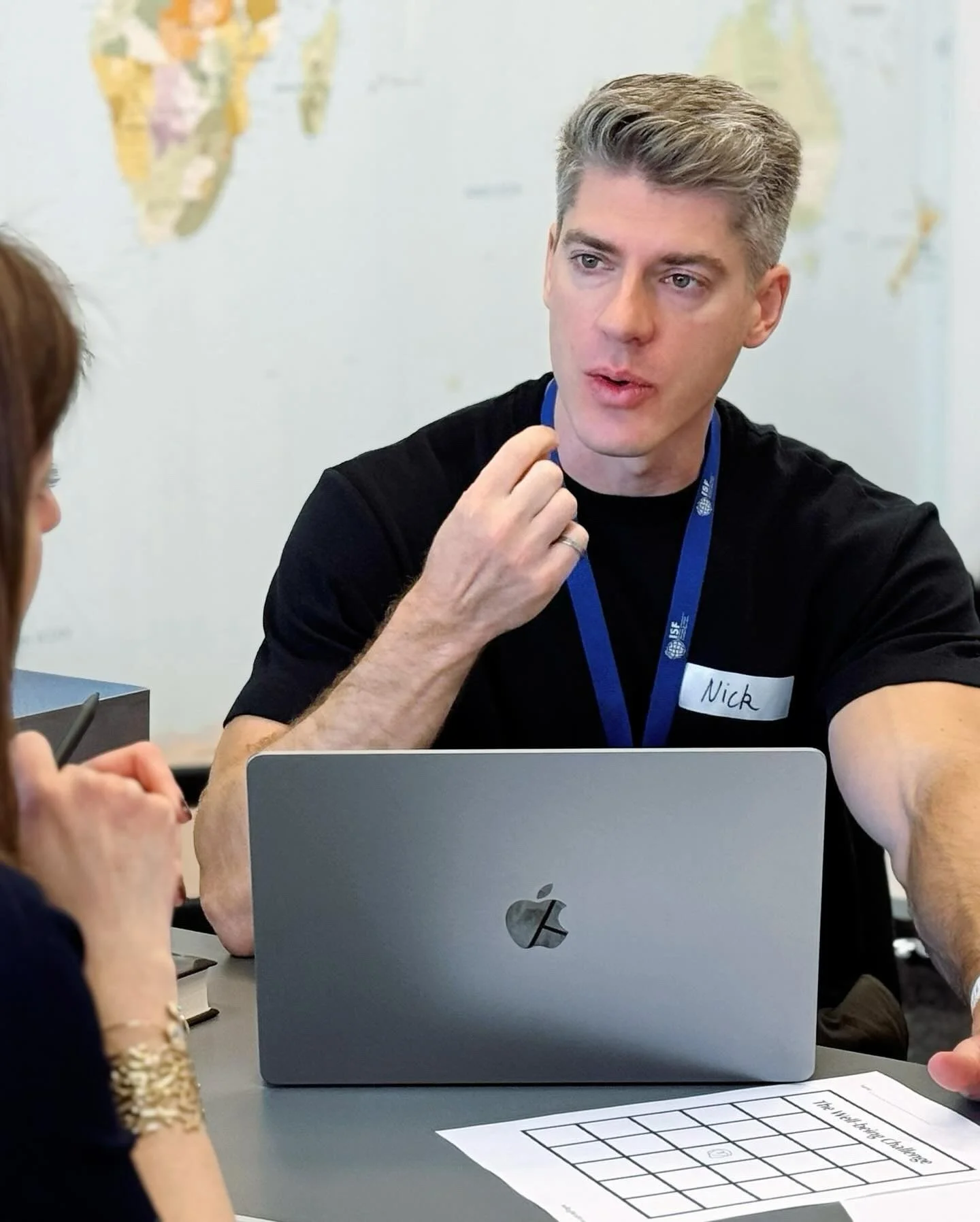 Nick, wearing a lanyard and name tag, sitting at a table talking while using a laptop during a school session.