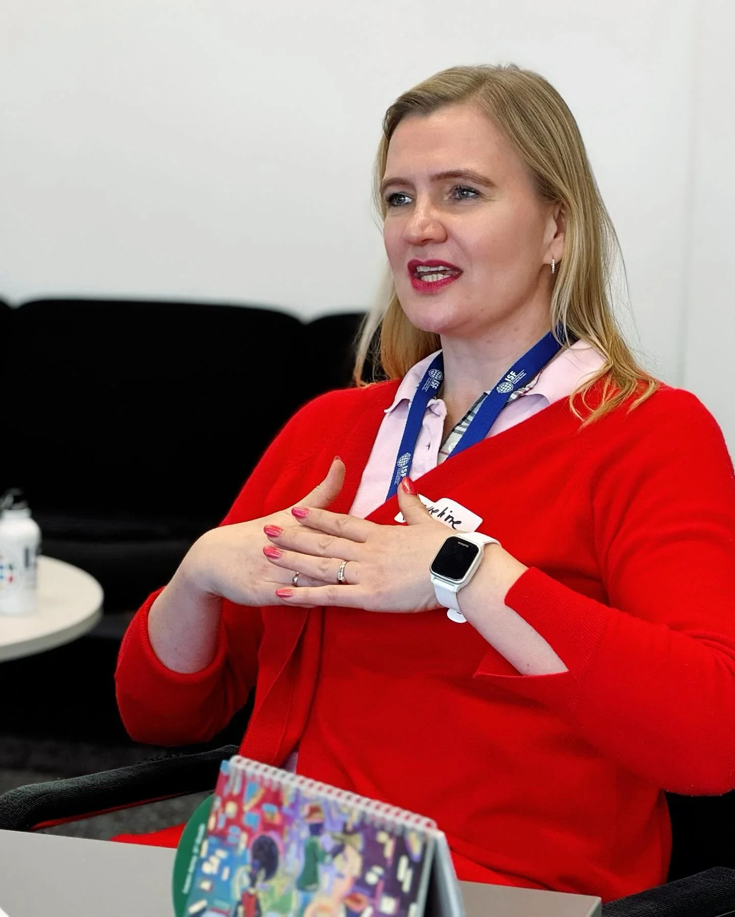 Jacqueline in a red cardigan and ID lanyard, sitting at a desk and gesturing while speaking.